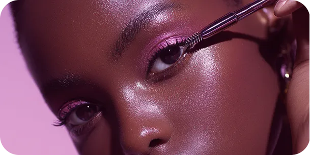 Close-up of a person applying mascara to their upper eyelashes with a makeup brush against a pink background.