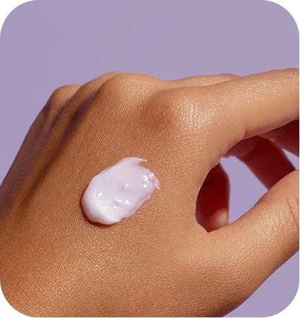 Close-up of a hand with a dollop of light pink cream on the back, against a lavender background.