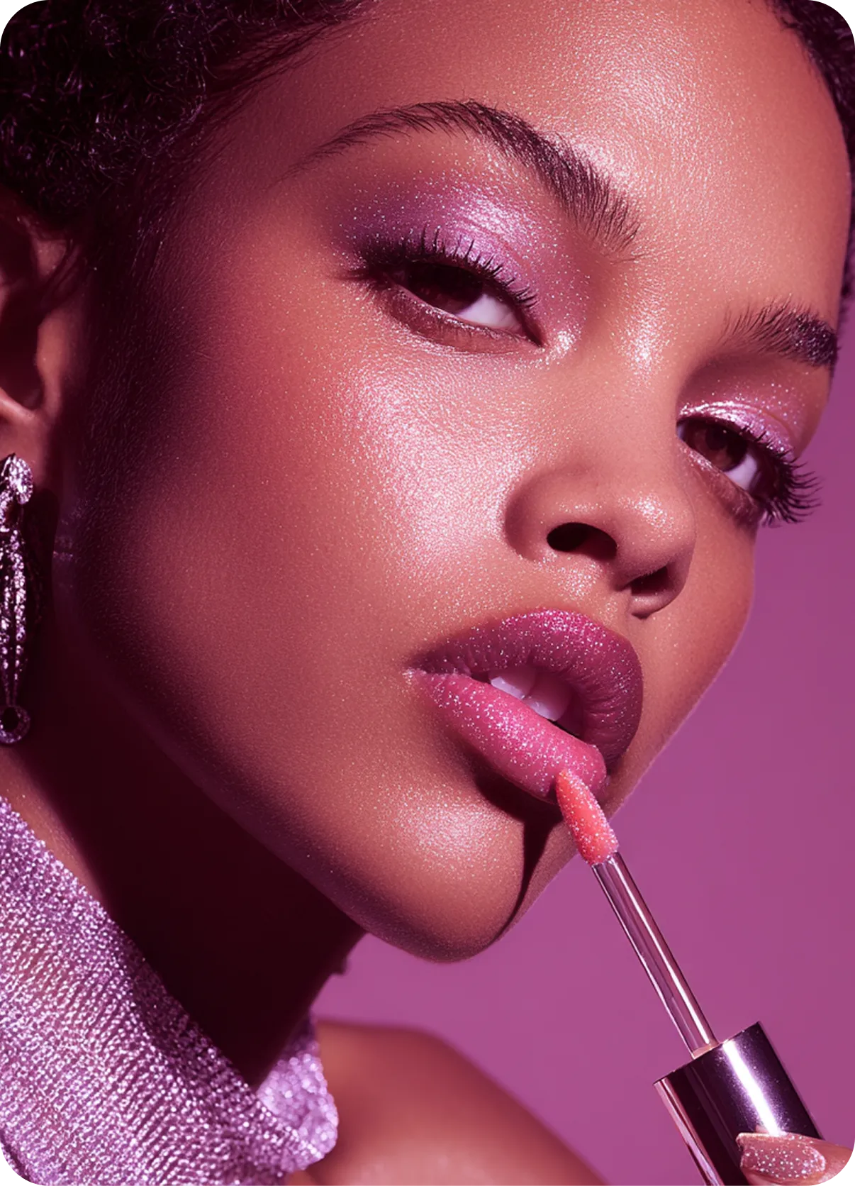 Close-up of a woman applying glossy pink lip gloss, wearing shimmering purple eyeshadow and glittering earrings against a purple background.