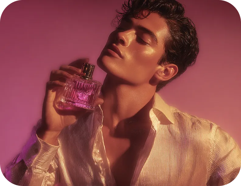 Man with glossy skin holds a pink perfume bottle close to his face against a pink background.