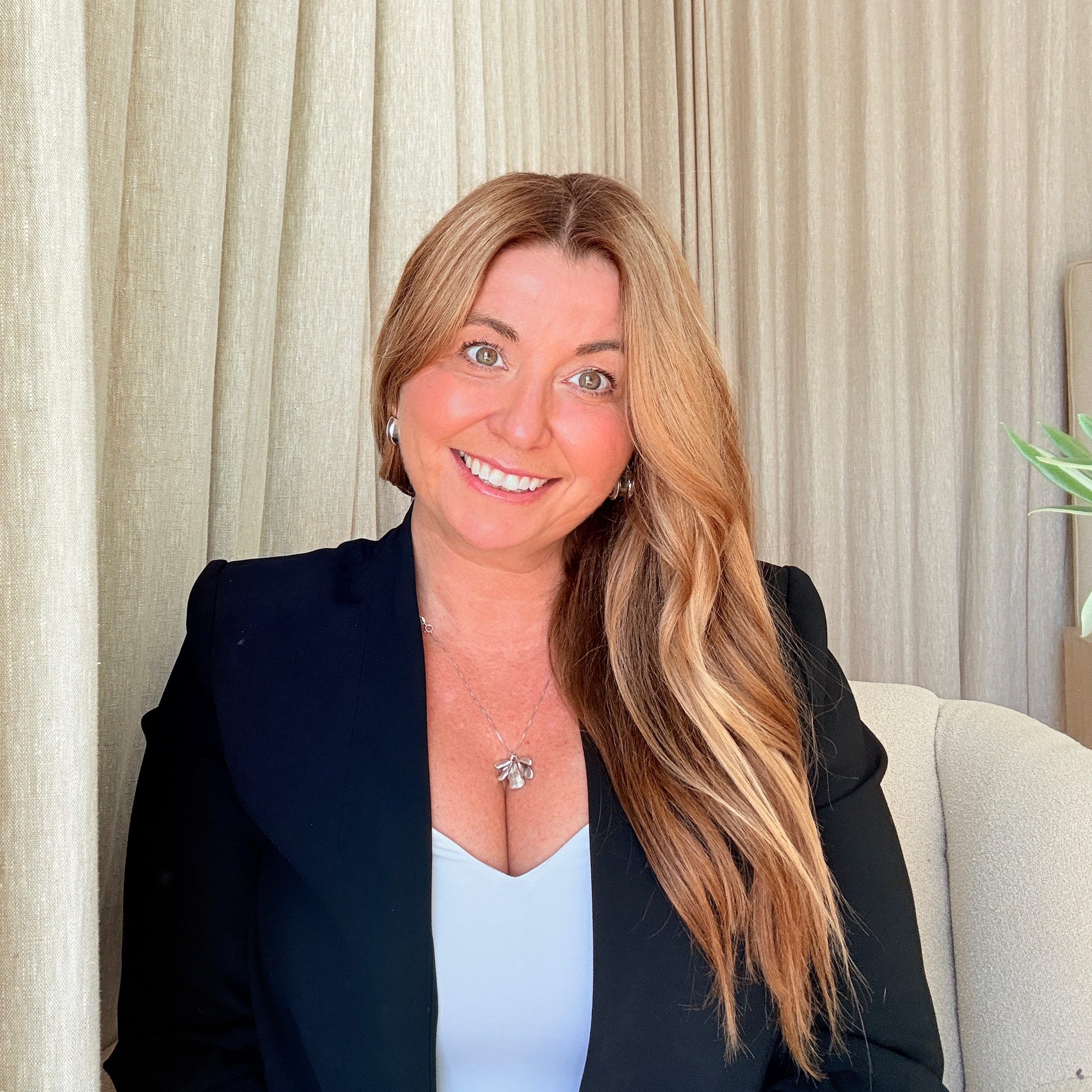 Smiling woman with long blonde hair wearing a black blazer and silver necklace, seated indoors in front of beige curtains.