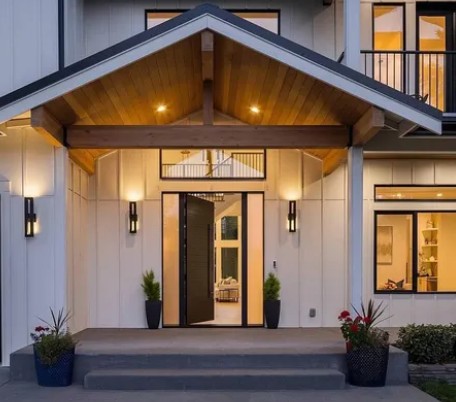 Modern farmhouse entry featuring smooth light-colored siding with clean vertical lines, a covered gabled porch with exposed wood beams, black trim accents, and large windows that highlight the home’s contemporary architectural design.