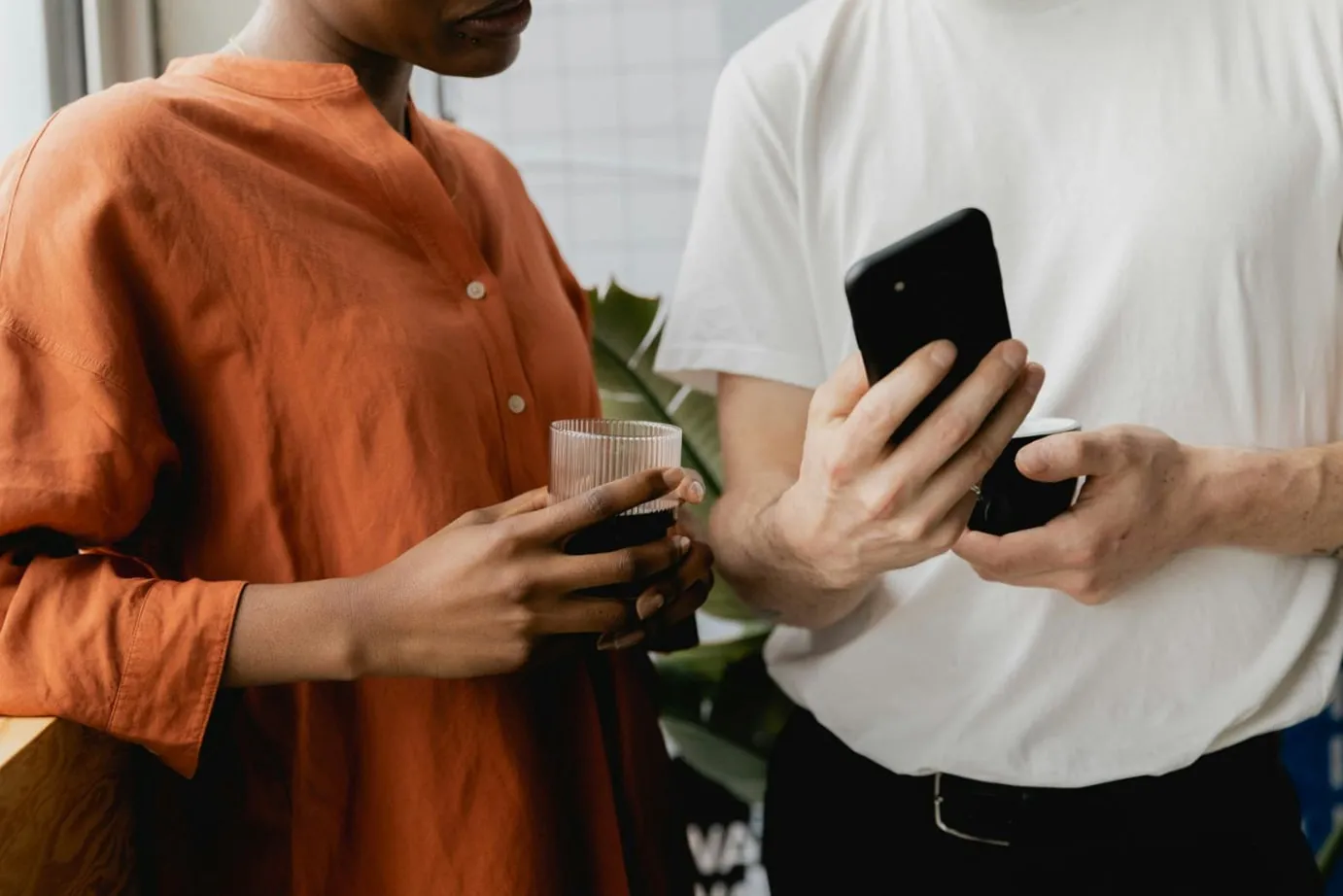 Two people holding drinks while looking at a phone.
