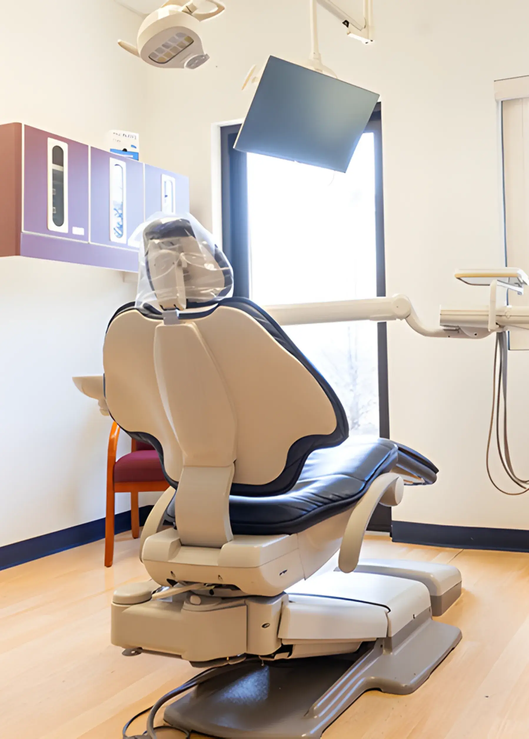 An empty dental chair in a bright office, equipped with an overhead light and monitor.
