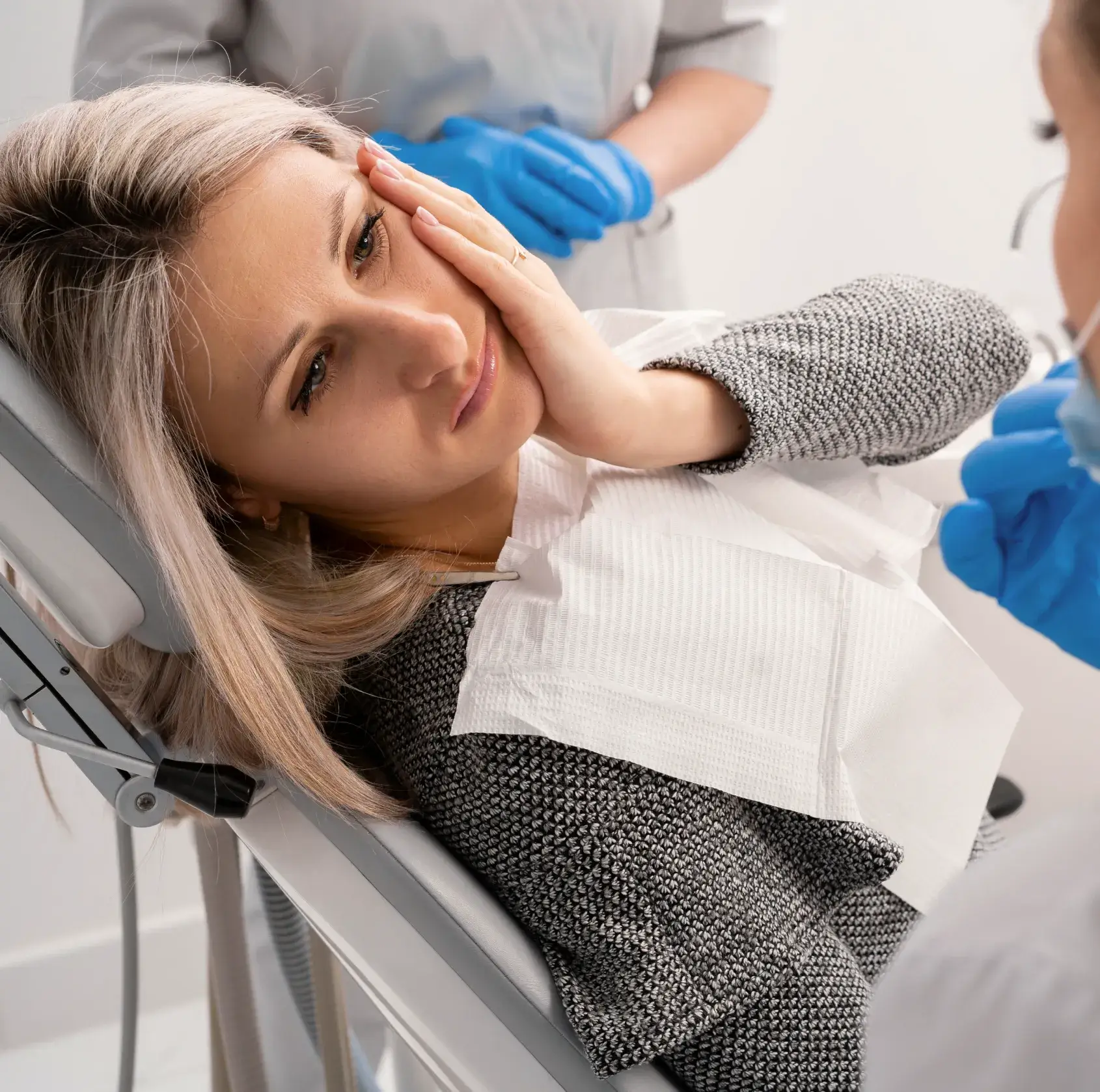 A woman sits in a dental chair, touching her cheek, while a dentist prepares to examine her.