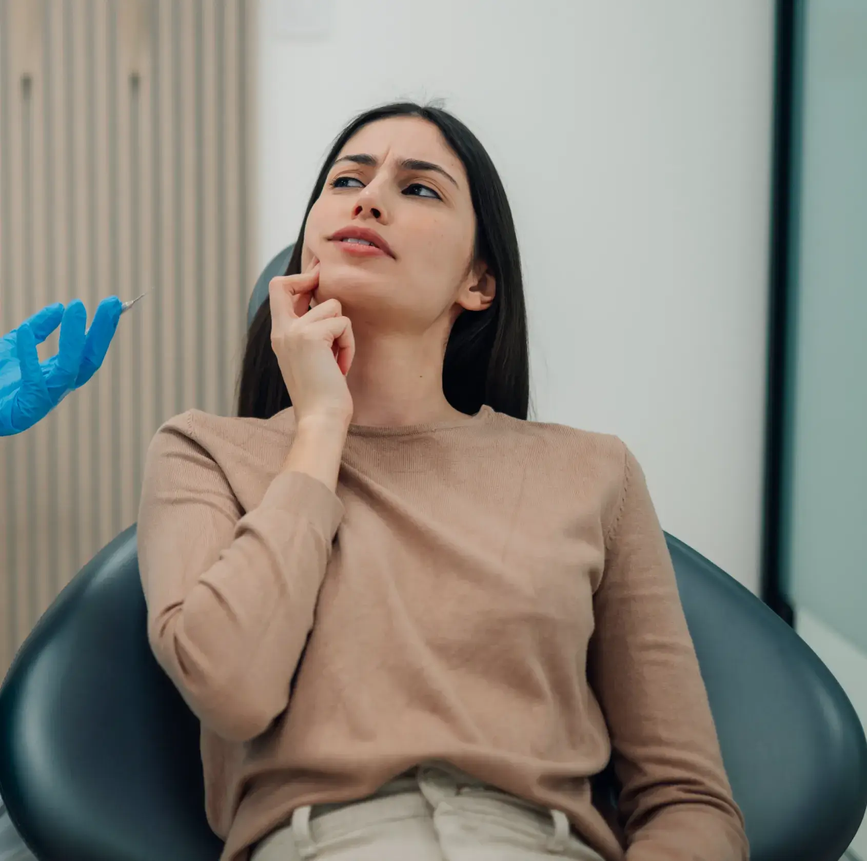 A woman sits in a dental chair, looking concerned, while a dentist holds a dental tool.