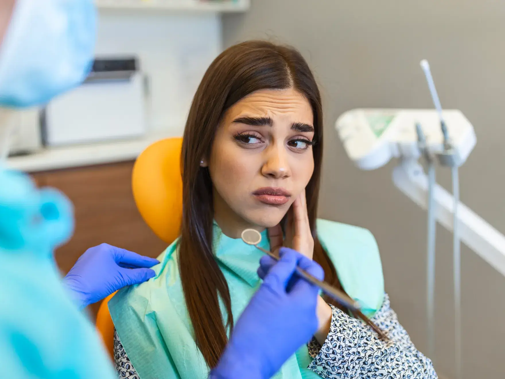 A woman sits in a dental chair, looking concerned, while a dentist holds tools near her face.
