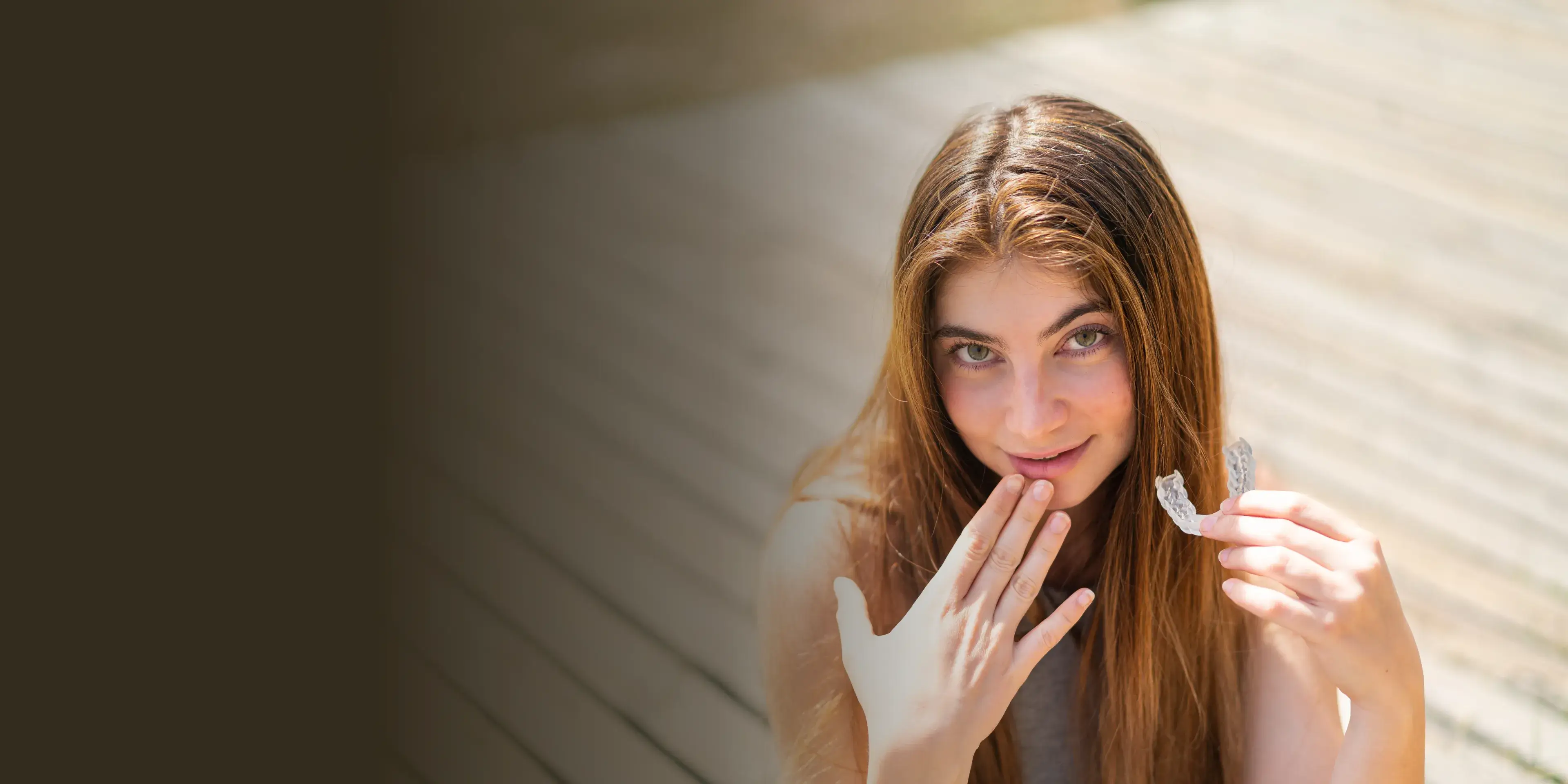 A woman with long hair holds a dental aligner and smiles, sitting on a wooden floor.