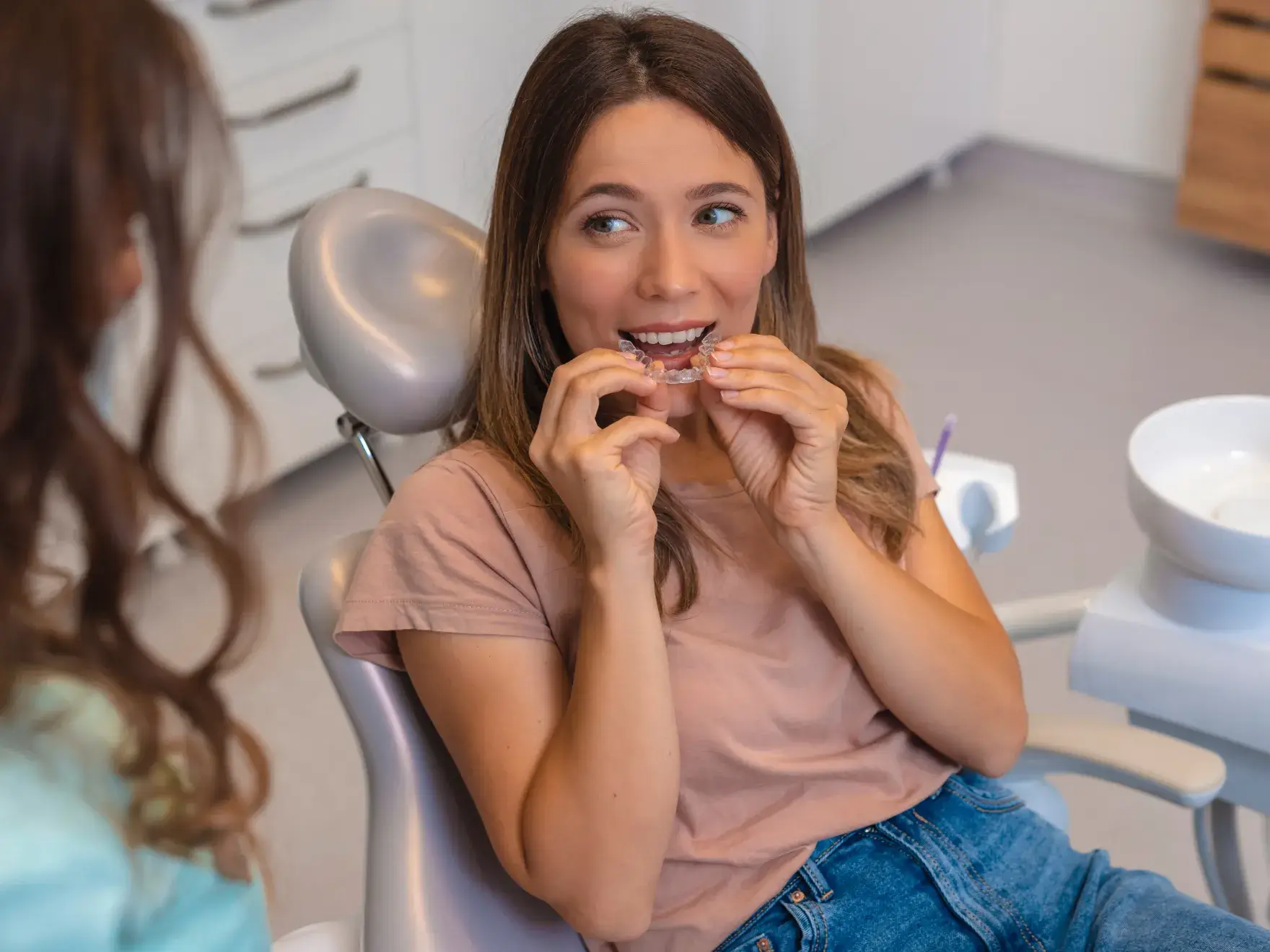 A woman sitting in a dental chair holds a clear aligner near her teeth, smiling.
