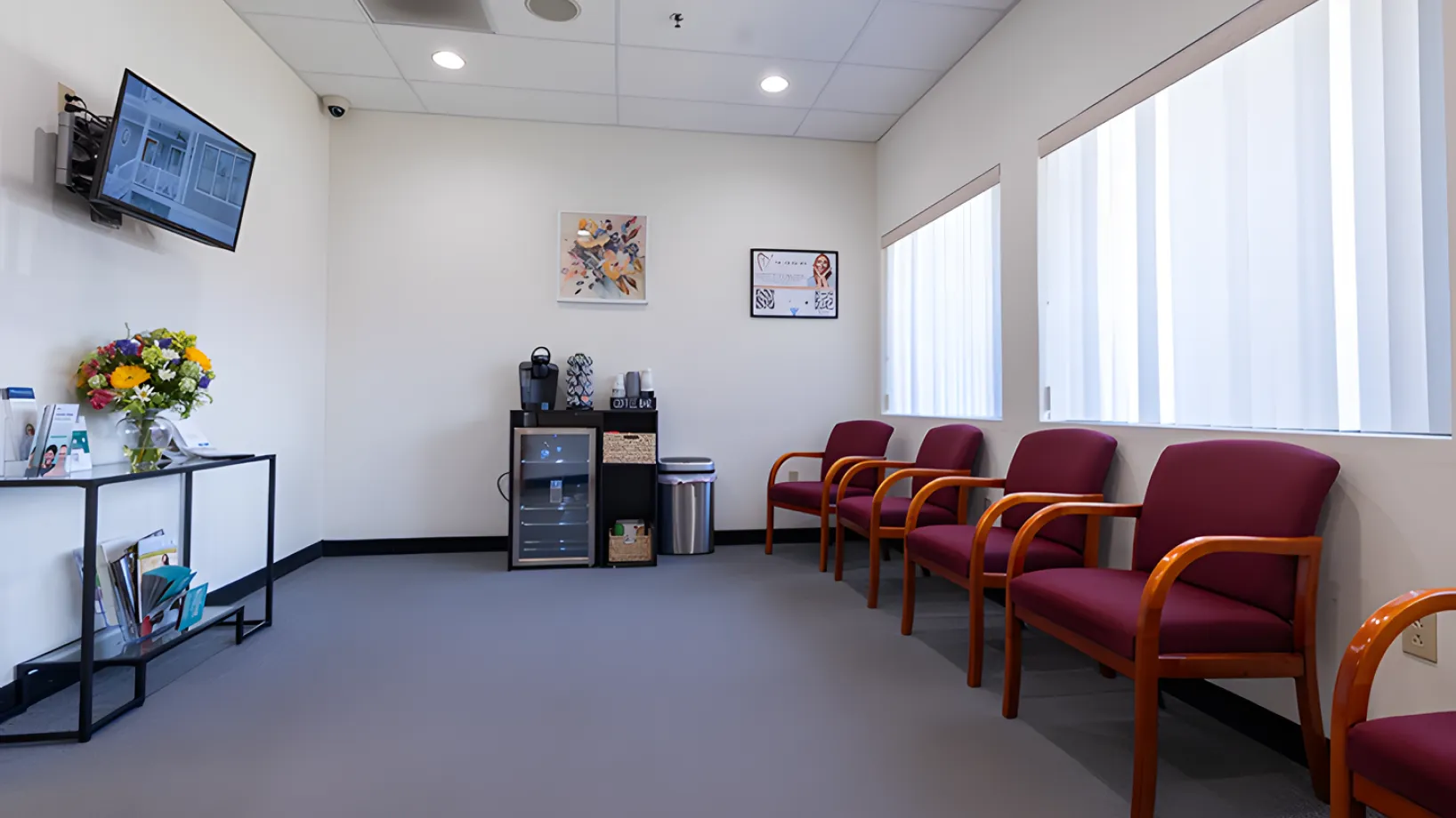 Five red chairs line the side of a small, tidy waiting room with a TV on the wall.