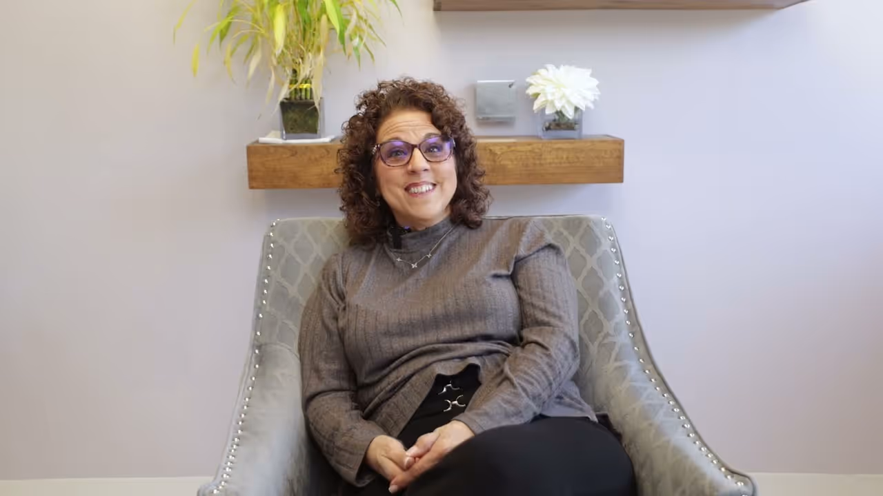 A woman with curly hair and glasses sits in a chair, smiling comfortably.