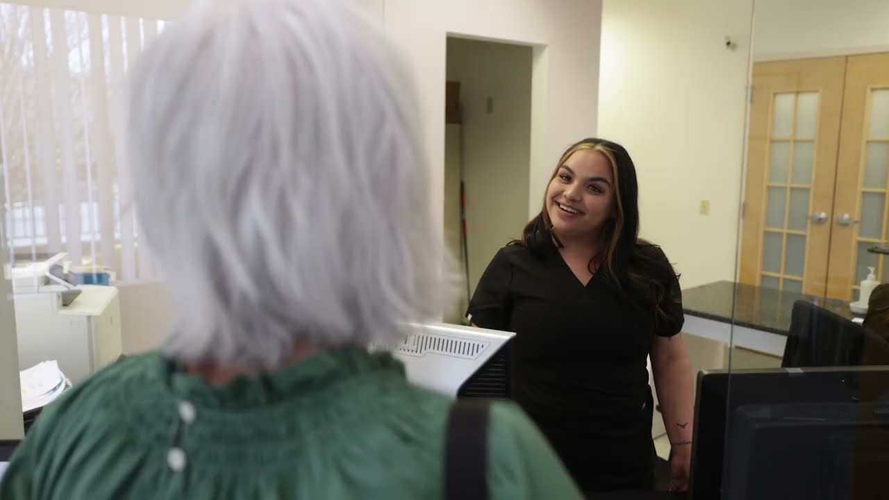A woman at a reception desk smiles and speaks to a visitor with white hair.