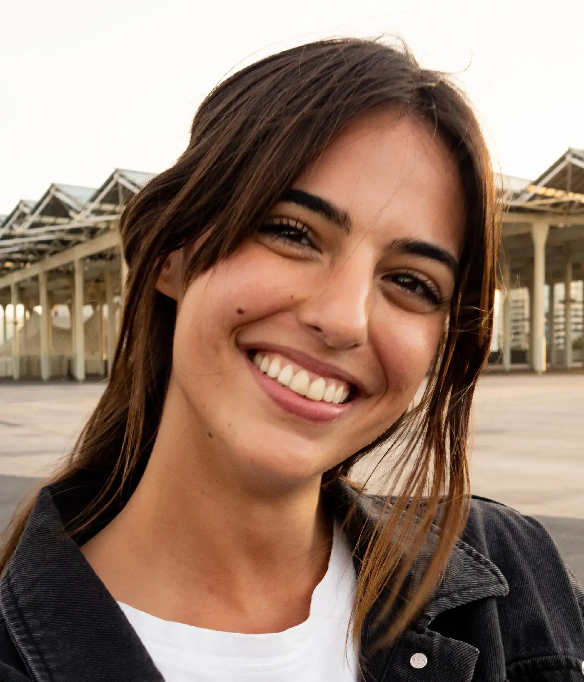 Smiling person with long hair in a black jacket stands outdoors near a row of structures.