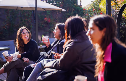 A small group of people enjoy casual conversation and coffee in a sunny outdoor garden area.
