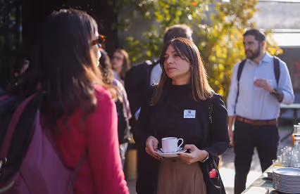 Two women talk during a coffee break at an outdoor networking event, one holding a cup and saucer.