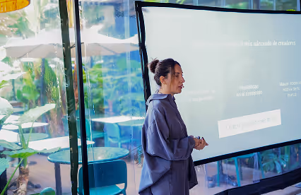A speaker presents next to a projected slide in a bright, glass-enclosed venue surrounded by lush greenery.