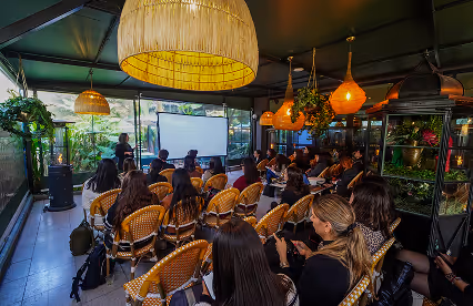A speaker presents to a group seated in a cozy, plant-filled semi-outdoor venue with warm hanging lights.