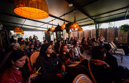 Attendees listen attentively during a presentation in a cozy, covered outdoor space with hanging lights.