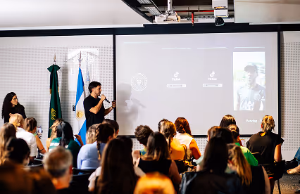 A presenter speaks in front of a large screen during an indoor conference, with an audience watching closely.