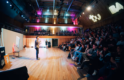 A speaker addresses a large crowd in a modern auditorium with dynamic lighting and tiered seating.
