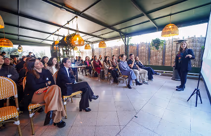 A speaker presents to a seated audience in a semi-outdoor venue with warm lighting and rattan chairs.