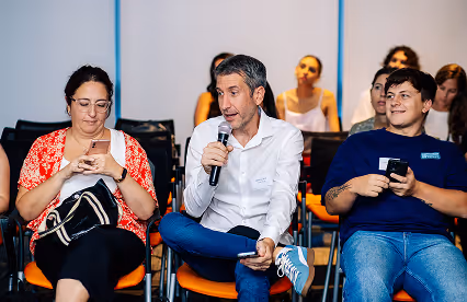 An audience member holds a microphone to ask a question while others engage with their phones or smile.