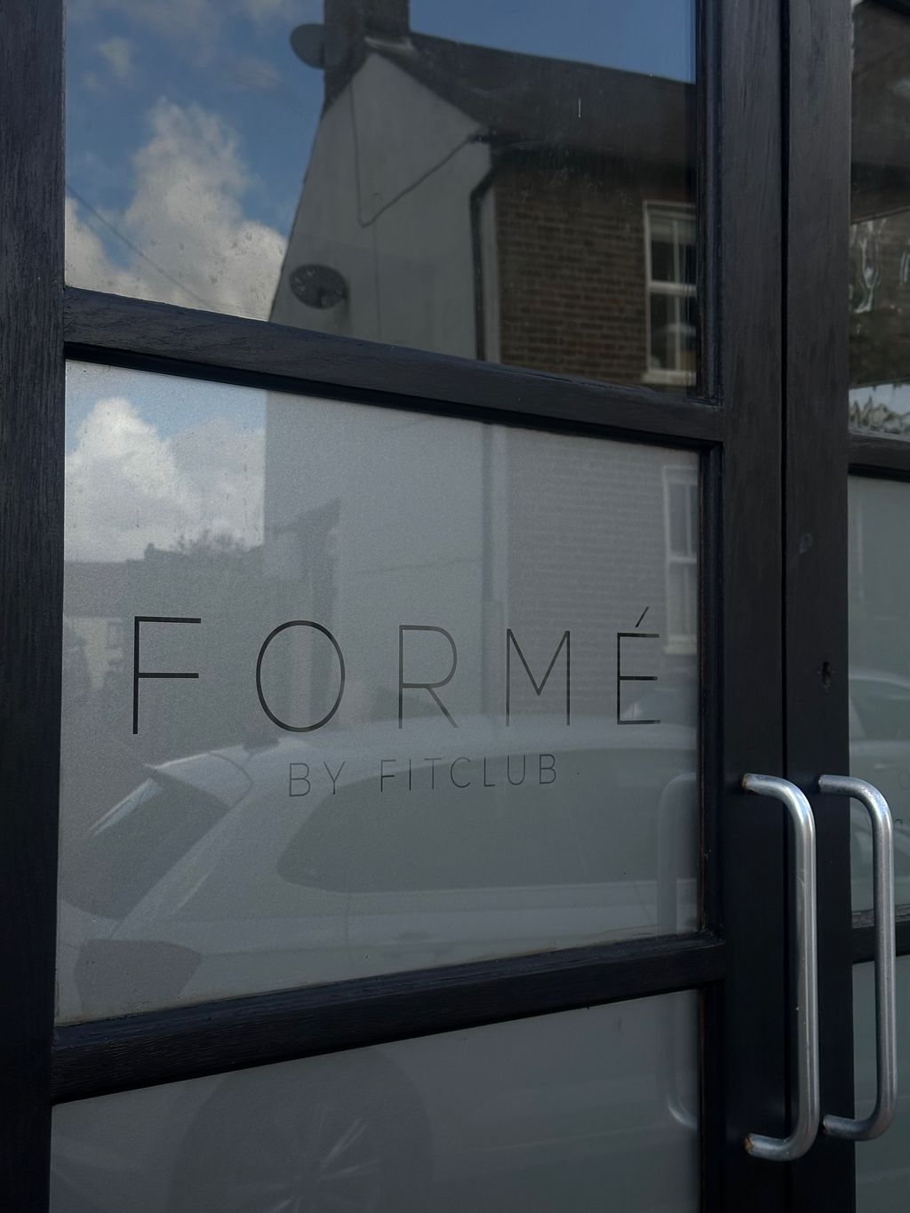 Glass door with black frame displaying the text 'FORMÉ BY FITCLUB' with reflections of nearby buildings and a car.