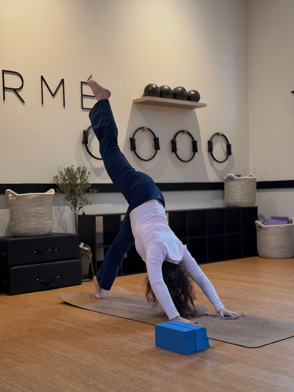 Person performing a yoga downward dog split pose on a mat with a blue yoga block in a studio.