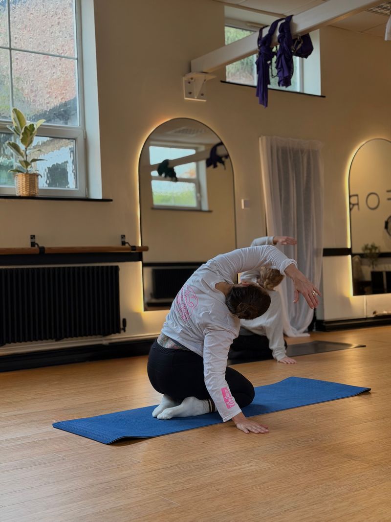 Two people practicing yoga stretches on mats in a bright studio with large arched mirrors and wooden floor.