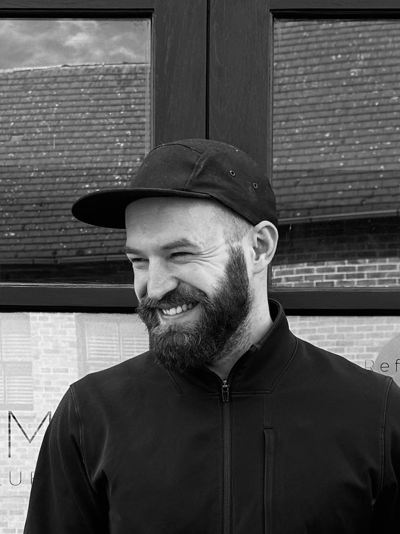 Smiling bearded man wearing a black cap and jacket standing in front of a window with reflections.