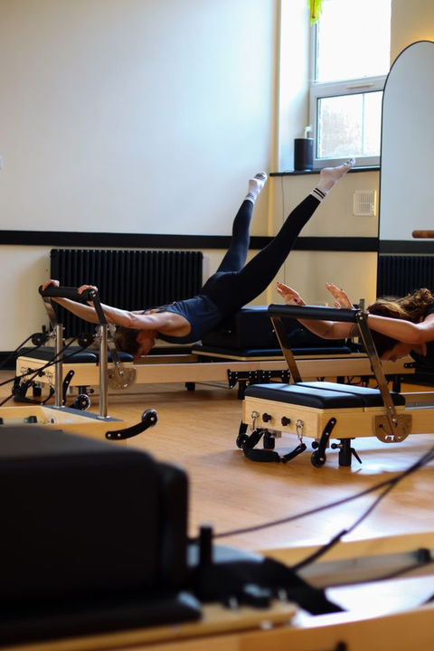 Person performing Pilates exercise on reformer machine with legs extended upward in a bright studio.
