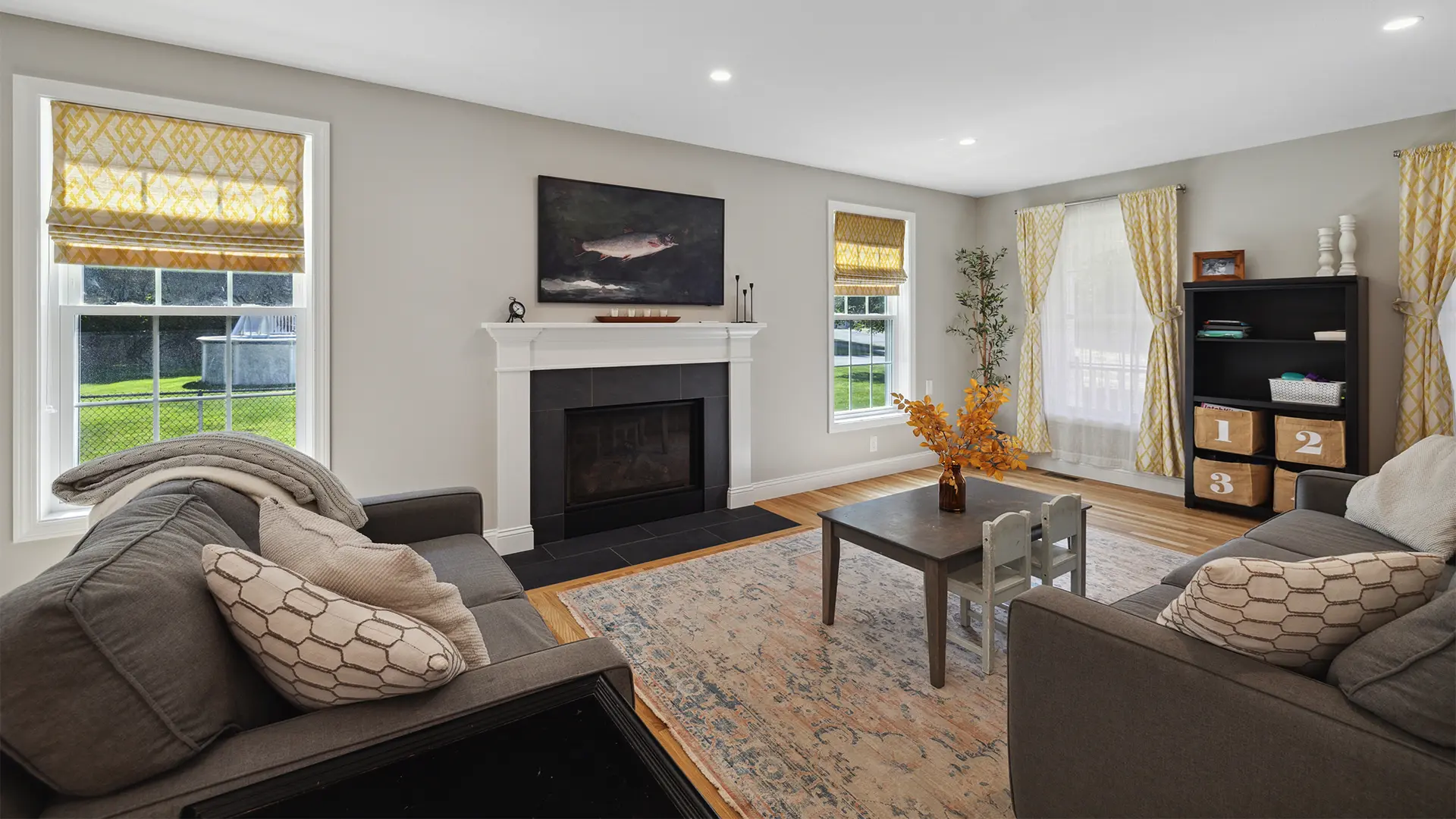 Bright modern living room with gray sofas, yellow patterned curtains, fireplace, and children’s play table.