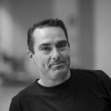 Black and white portrait of a man with short dark hair wearing a dark shirt, smiling slightly.