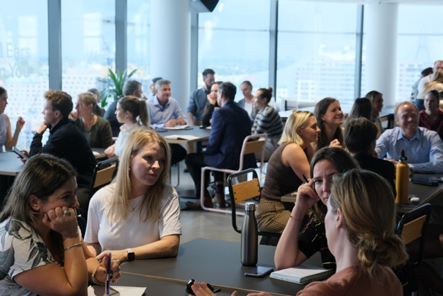 Groups of people engaged in conversations and discussions around tables in a large modern office space with floor-to-ceiling windows.
