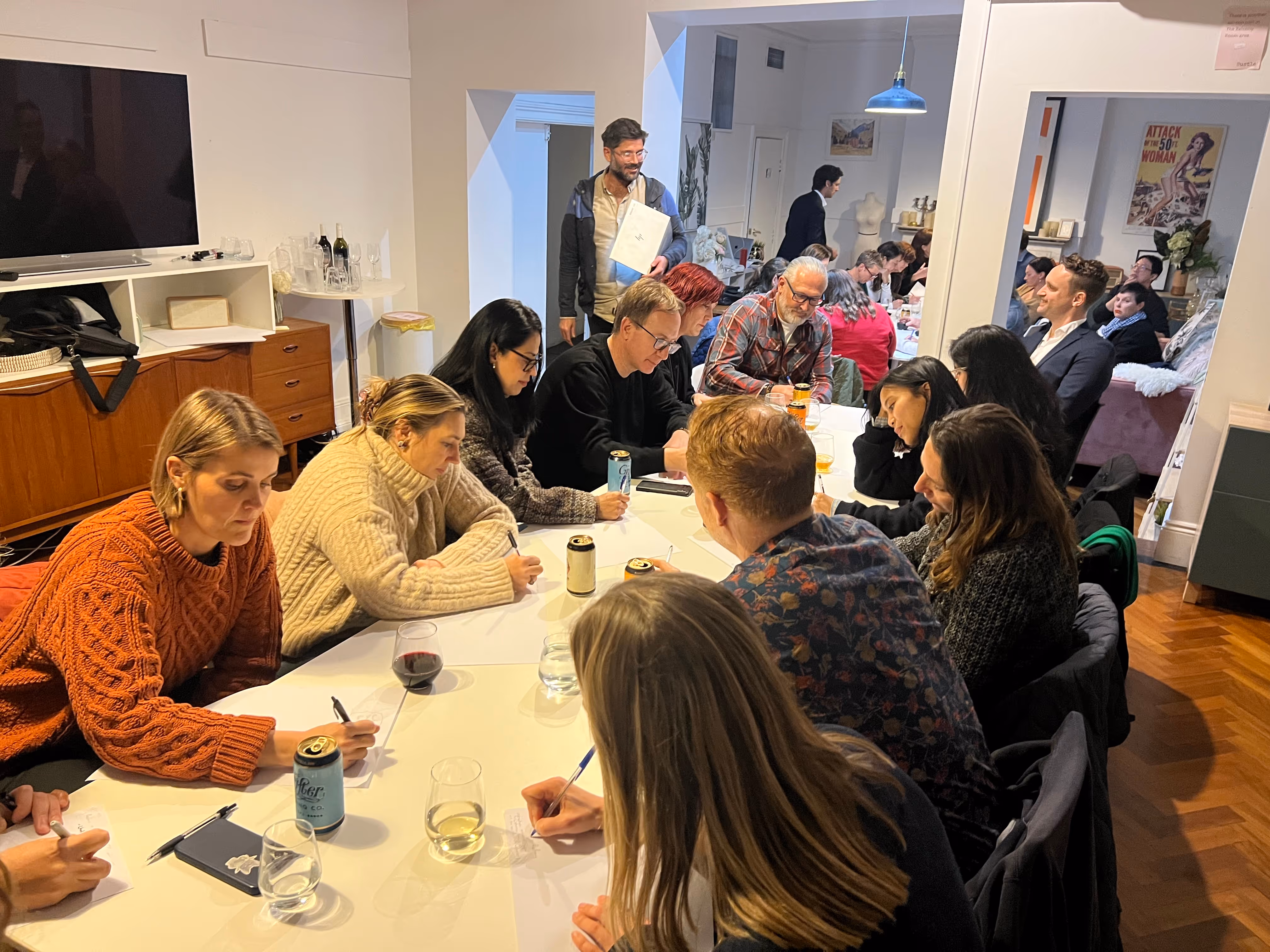 Group of people gathered around a long white table, some writing on papers, in a cozy room with wood flooring and modern decor.