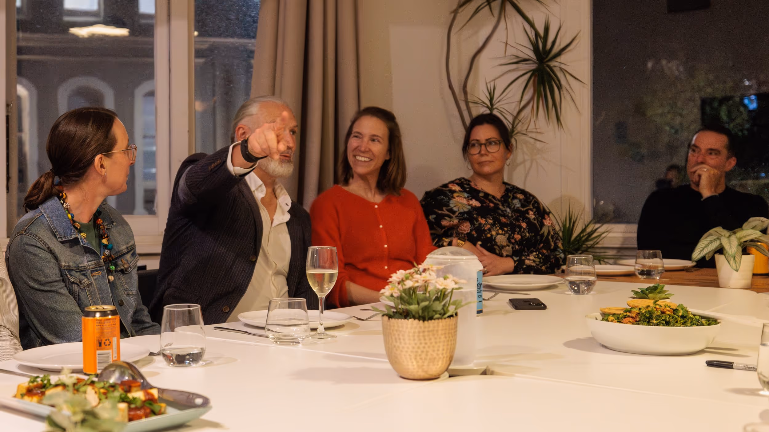 Five people seated around a dinner table engaging in conversation, with plates, glasses, and plants on the table.