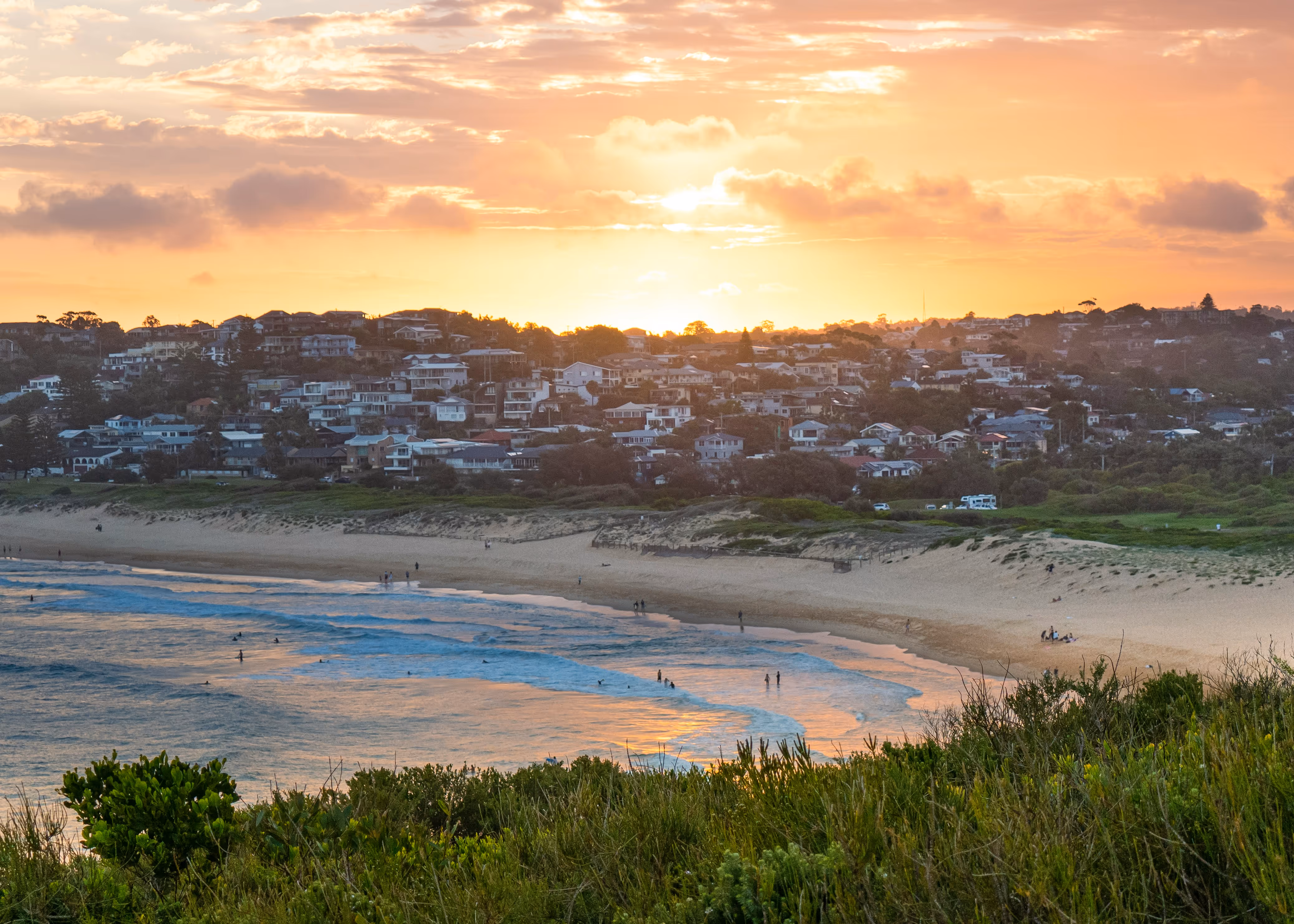 Sunset over a coastal town with sandy beach, ocean waves, and people in the water and on the shore.