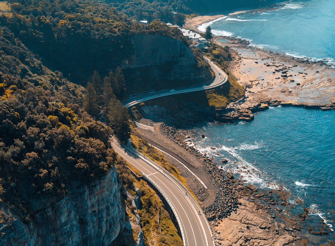Coastal road winding around a rocky cliff beside the ocean with clear blue water and scattered rocks.