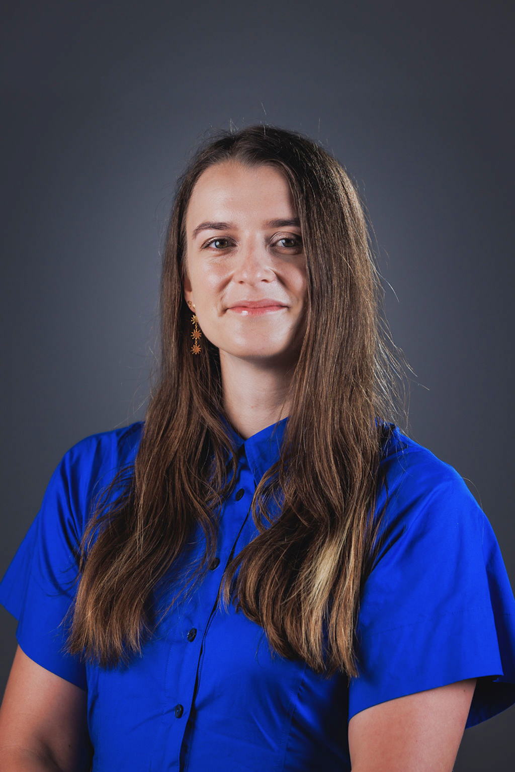 Woman with long brown hair wearing a blue button-up shirt and gold star-shaped earrings, smiling gently against a dark gray background.