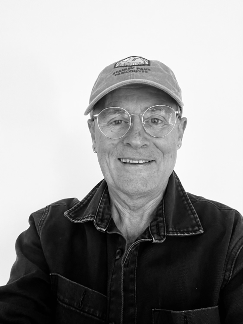 Smiling man wearing round glasses, a baseball cap, and a dark jacket against a white background.