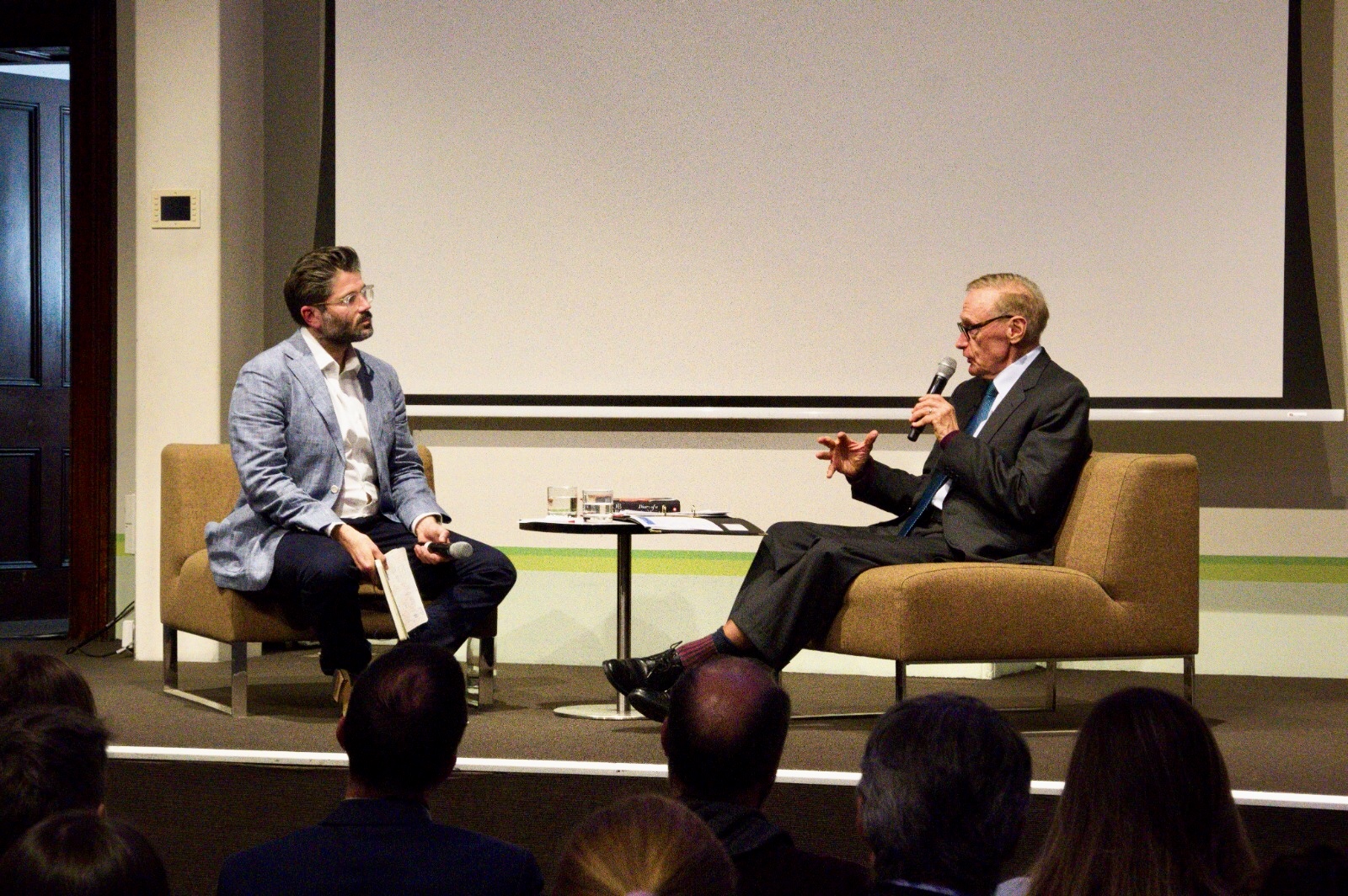 Two men seated on stage chairs engaged in a discussion, one holding a microphone and speaking while the other listens attentively, with a small table and audience in front.