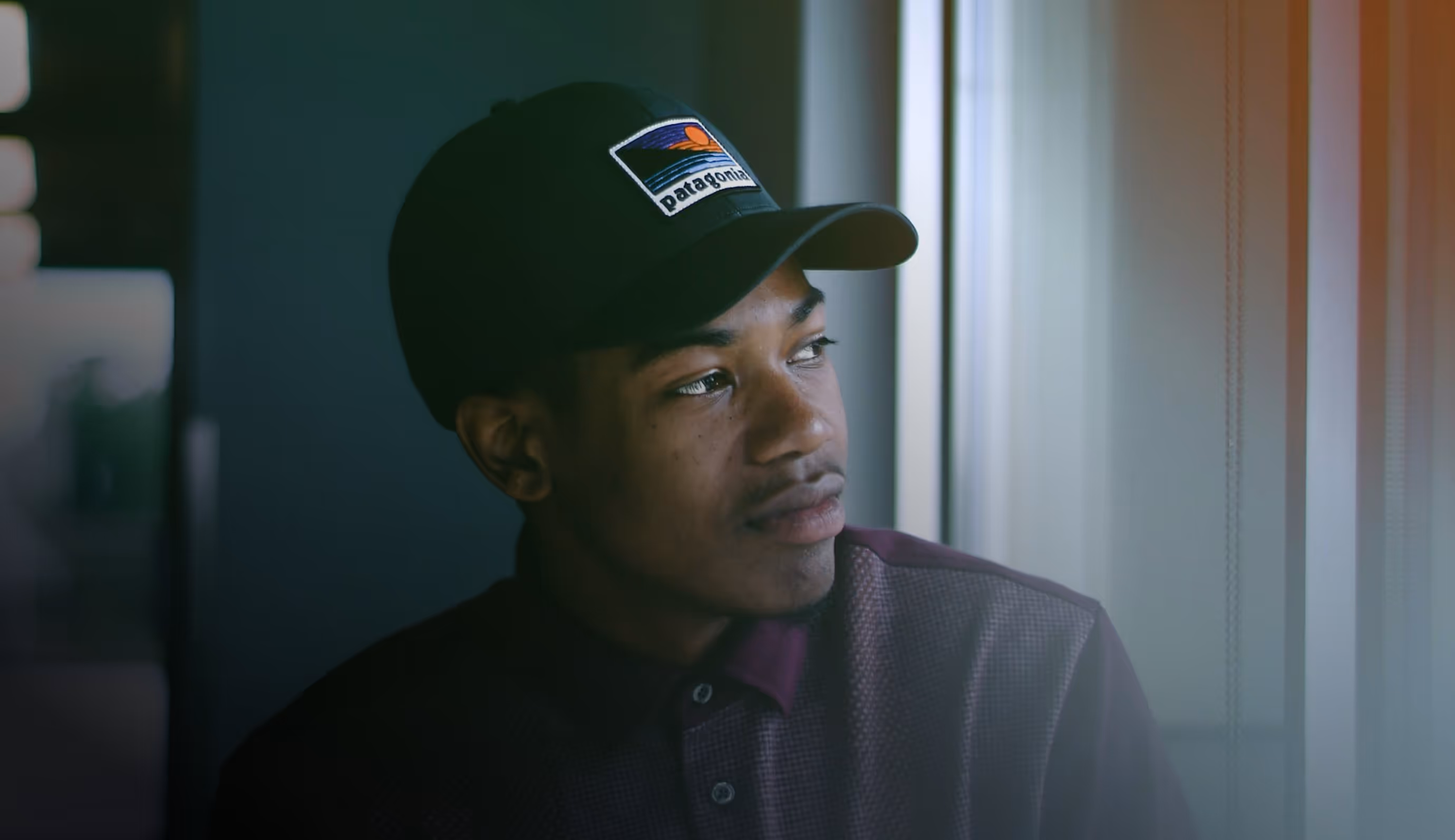 Photograph of a young man wearing a hat, sitting down and looking out a window