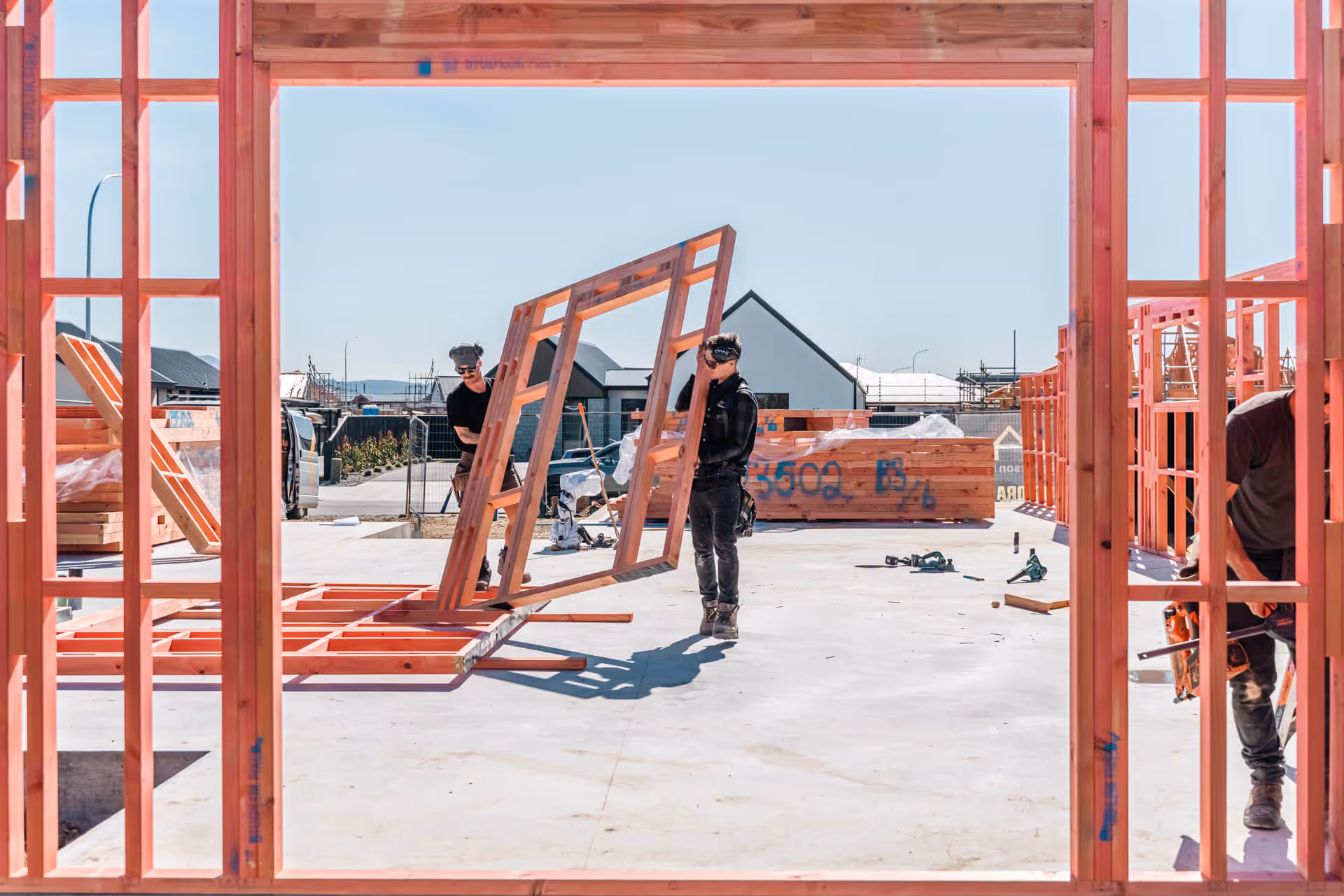 Two construction workers lifting a wooden frame inside a house under construction with exposed wooden studs and concrete floor.