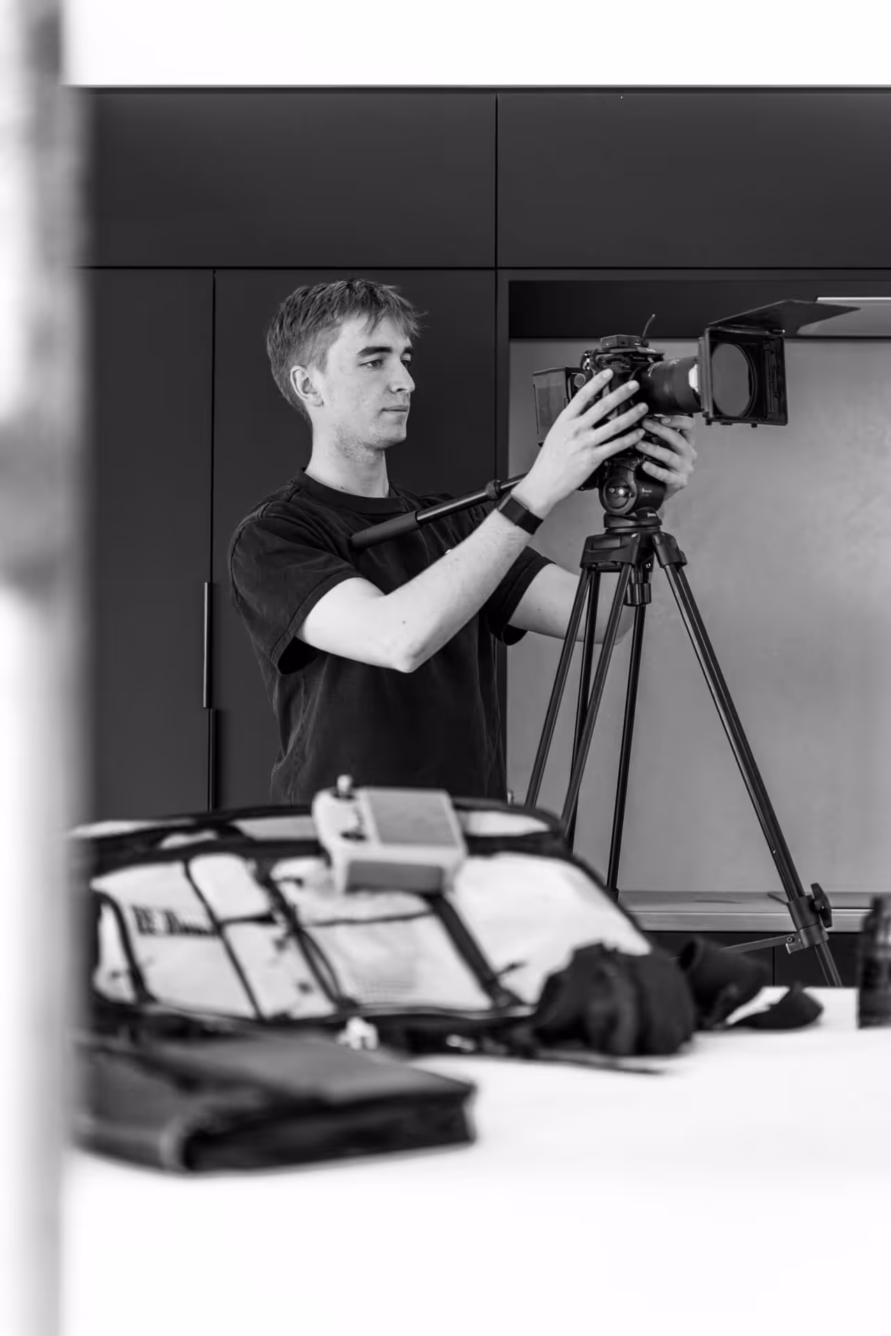 Man adjusting a professional camera on a tripod indoors with camera gear on a table in the foreground.