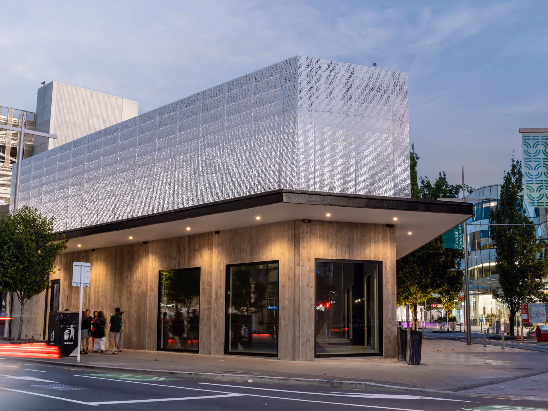 Modern two-story building with illuminated perforated upper facade and wooden lower walls on a city street at dusk.