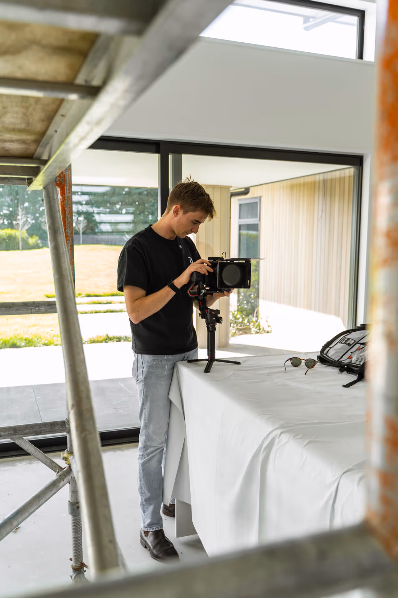 Man adjusting a professional camera mounted on a handheld stabilizer tripod on a table covered with white cloth indoors near large windows.