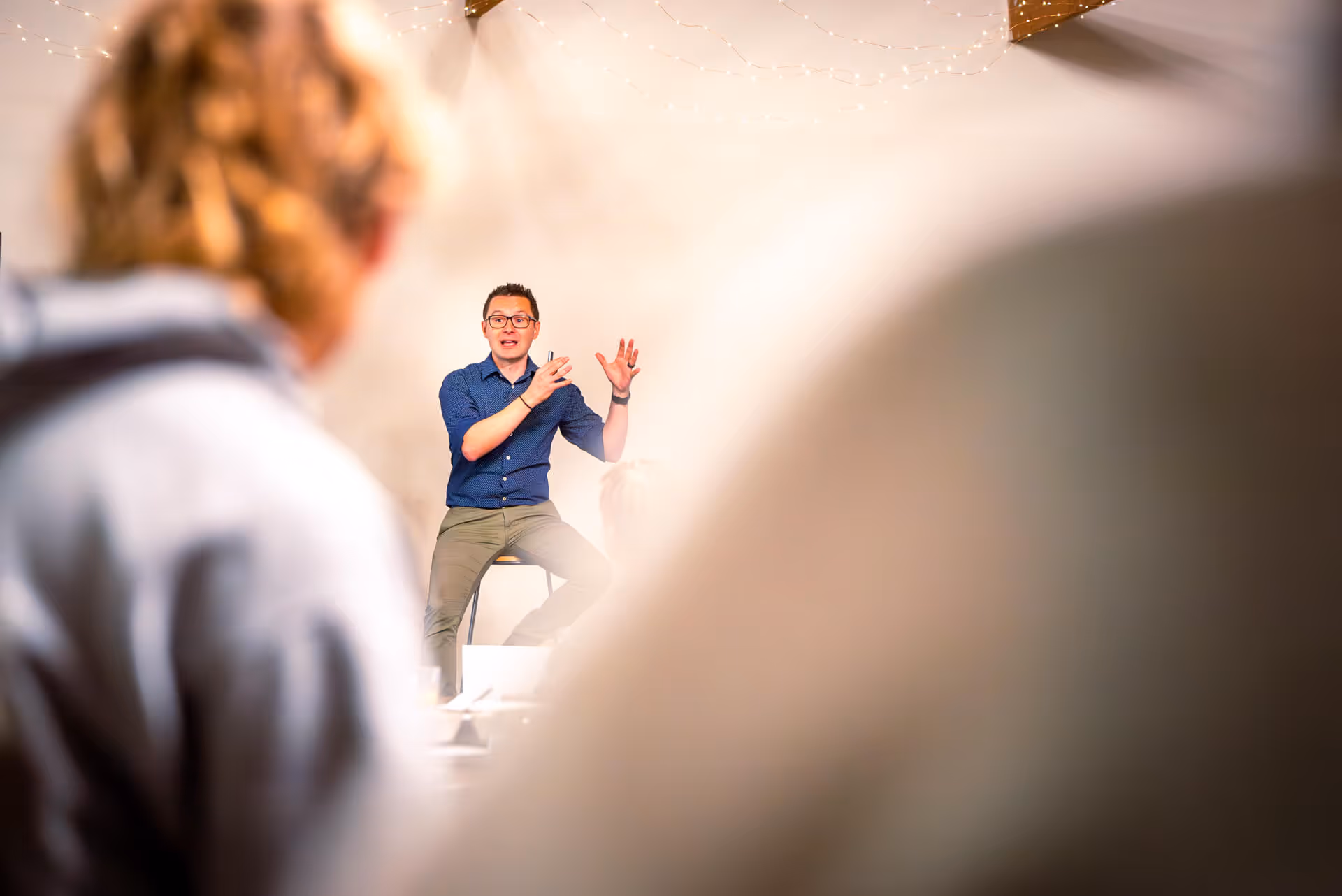 Man wearing glasses and blue shirt speaking and gesturing to an audience in a bright room.