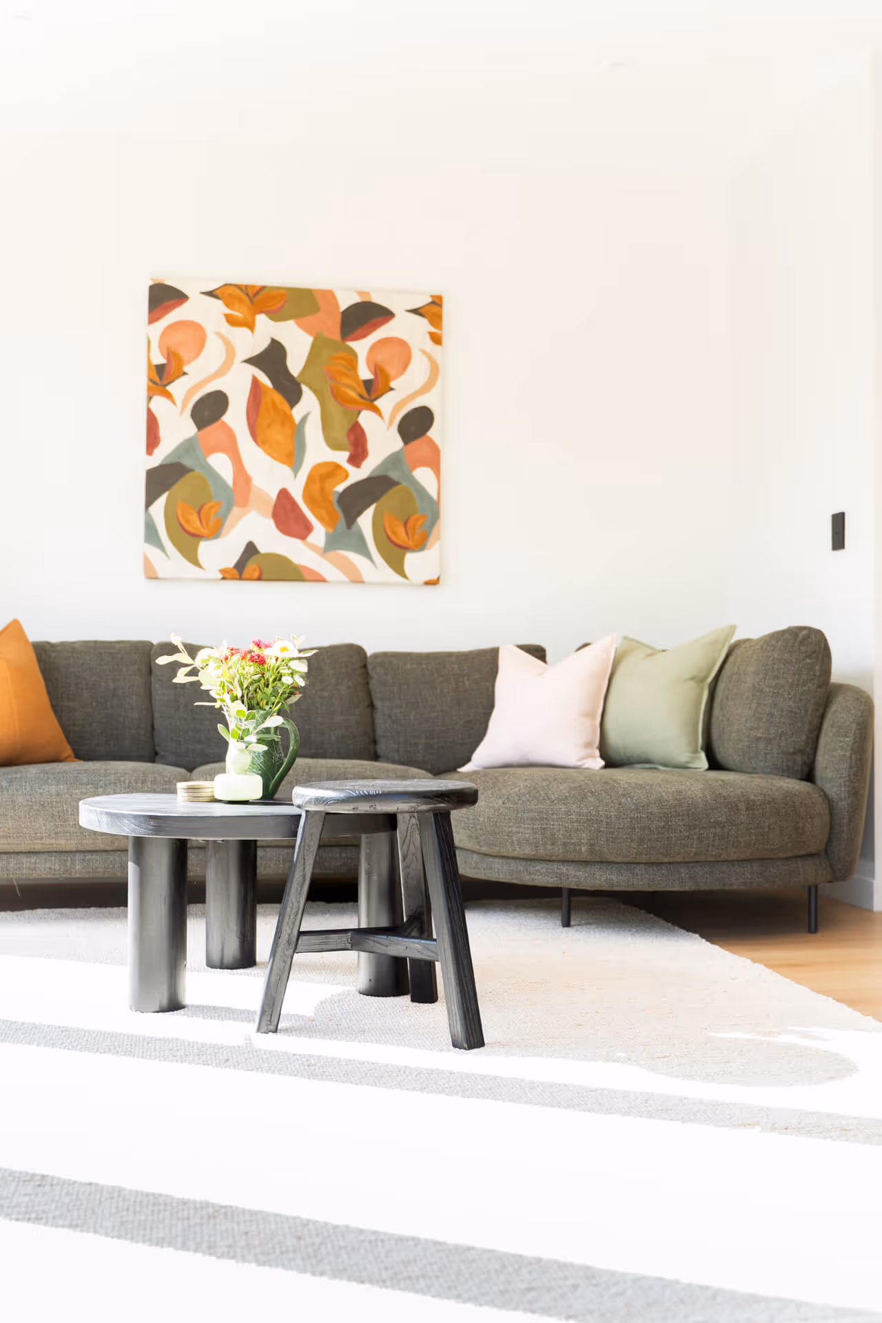 Modern living room with dark gray sofa, pastel and orange cushions, abstract leaf art on wall, black coffee table, and fresh flowers in vase.