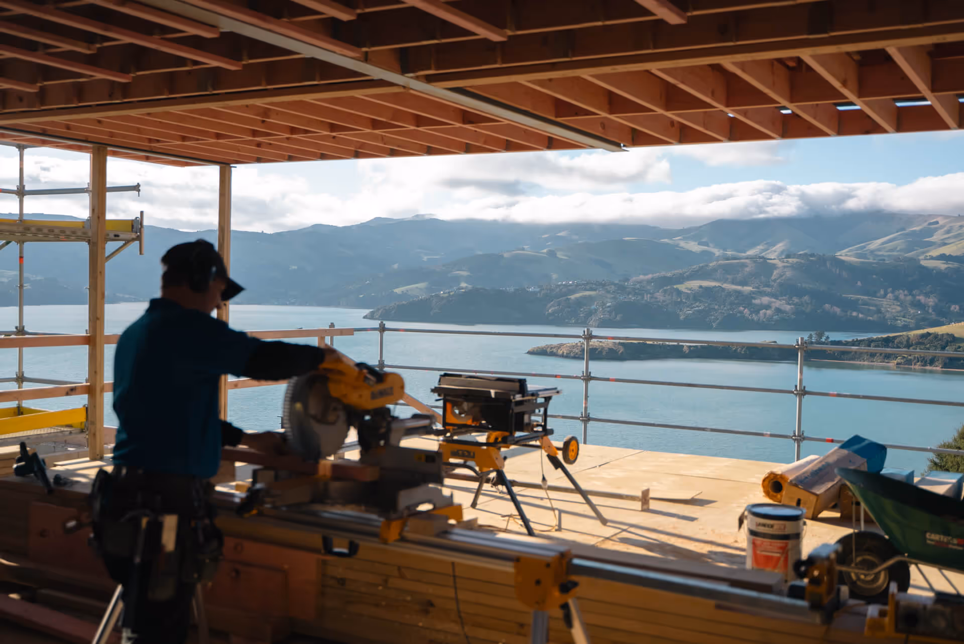 Construction worker using a power saw inside a wooden frame house with a scenic view of mountains and a lake through open walls.
