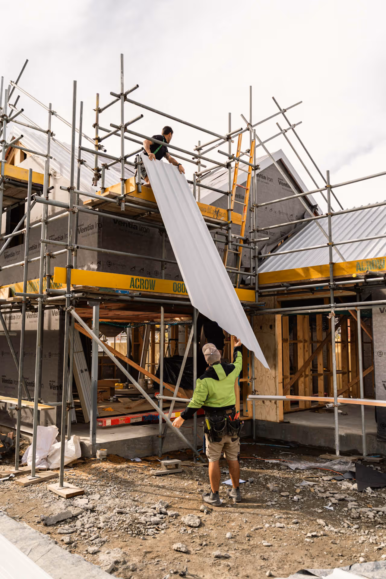 Two construction workers installing a white metal roofing sheet on scaffolding around a house under construction.